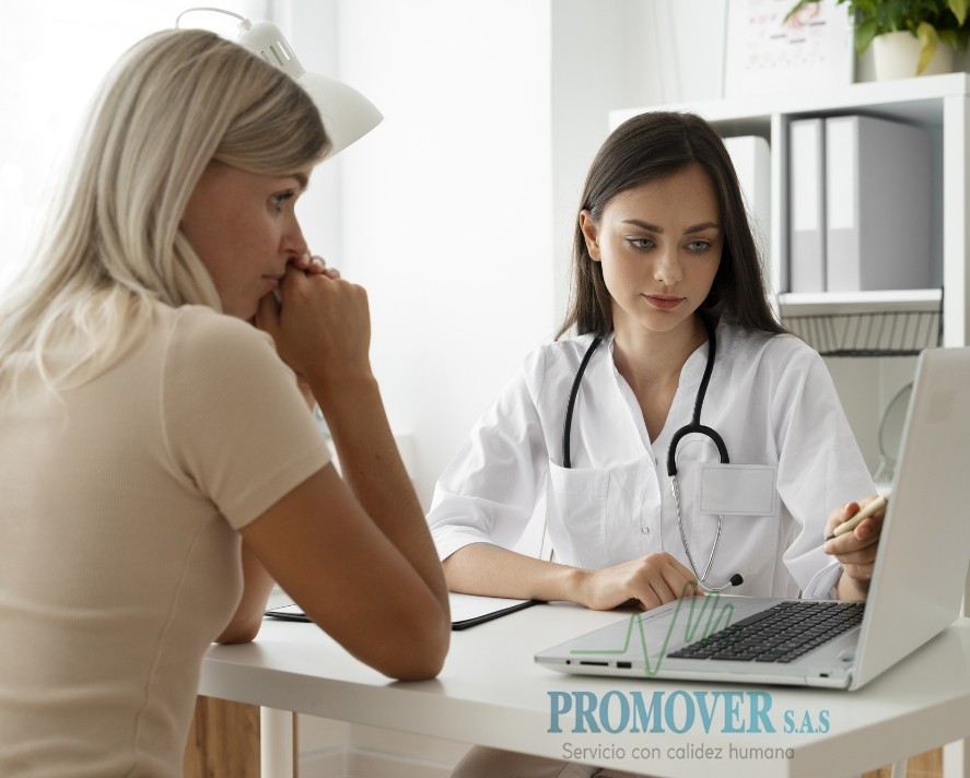 Médica de Promover S.A.S. atendiendo a una paciente en consulta, ambas sentadas frente a un computador portátil en un entorno clínico moderno y luminoso. El logo de la empresa aparece en la parte inferior con el eslogan Servicio con calidez humana